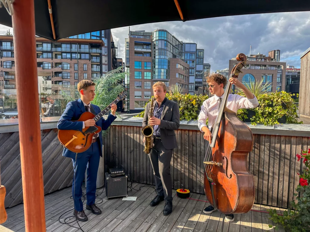 A jazz trio performing on a rooftop, featuring a guitarist, saxophonist, and double bass player, with modern buildings in the background.